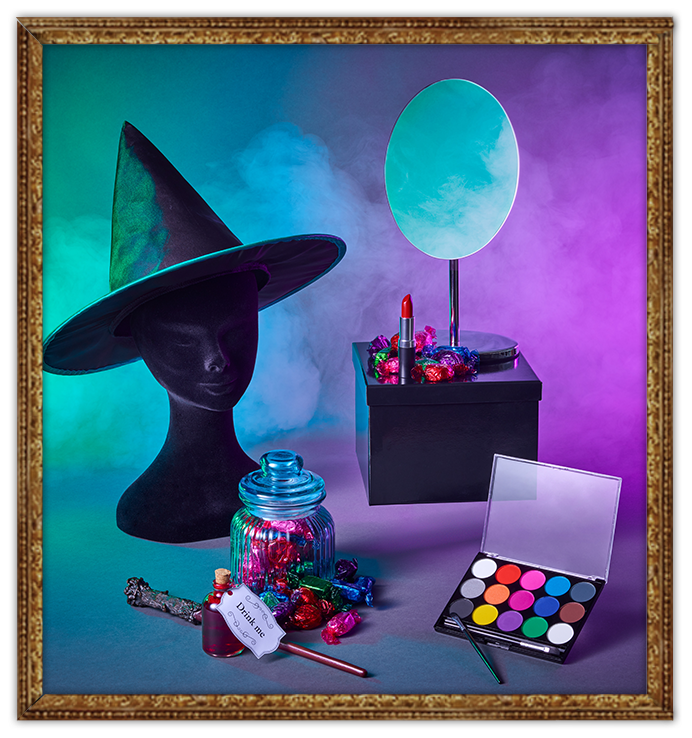 Black mannequin head with witch hat, jar of sweets, wand, makeup and a mirror, all shrouded in smoke